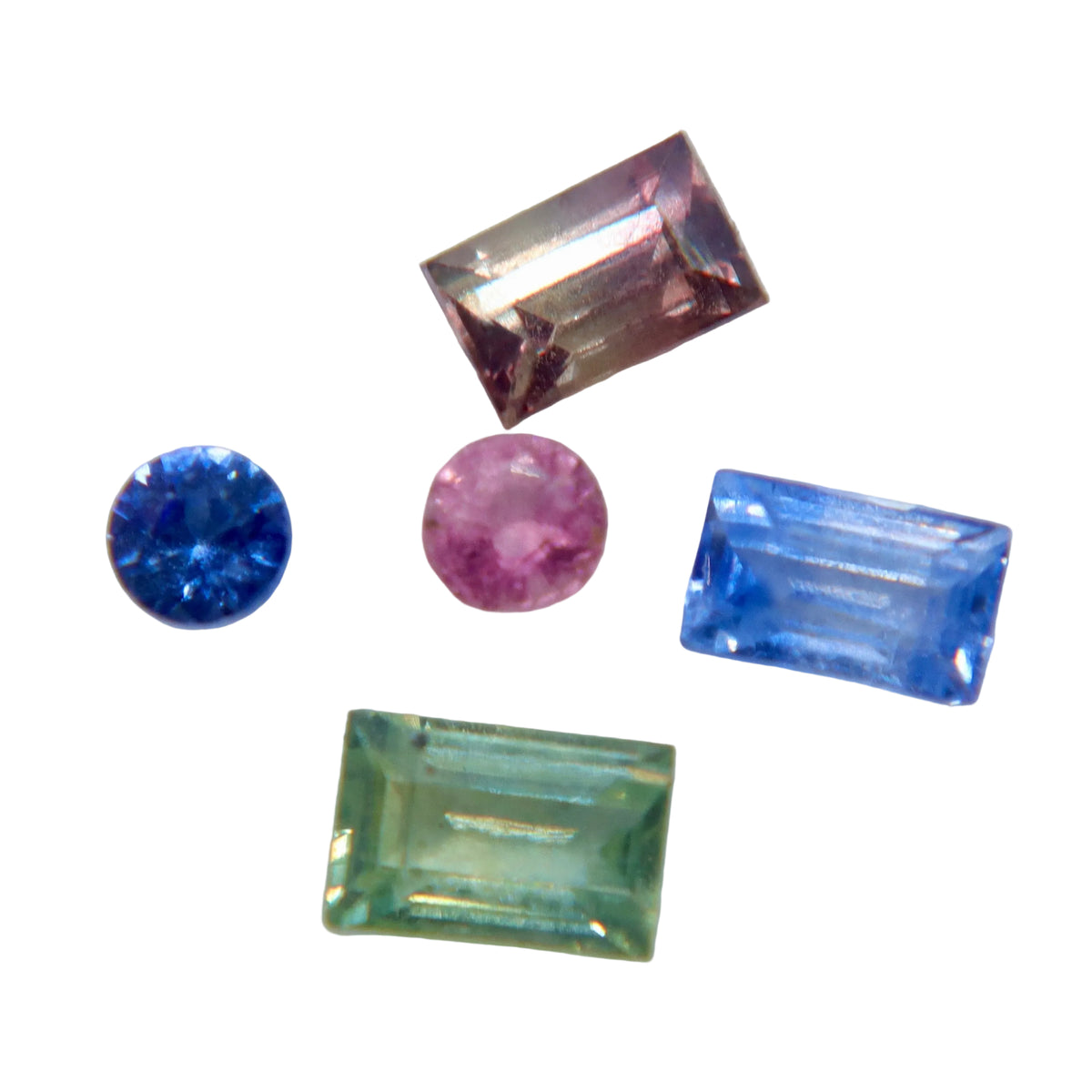 Five loose gemstones in different colors and shapes arranged on a light gray background: a rectangular pink stone, a round blue stone, a round pink stone, a rectangular light blue stone, and a rectangular light green stone.
