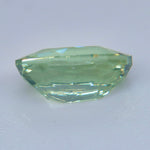A faceted light green gemstone with a rounded, cushion-like shape, displayed on a plain light gray background.