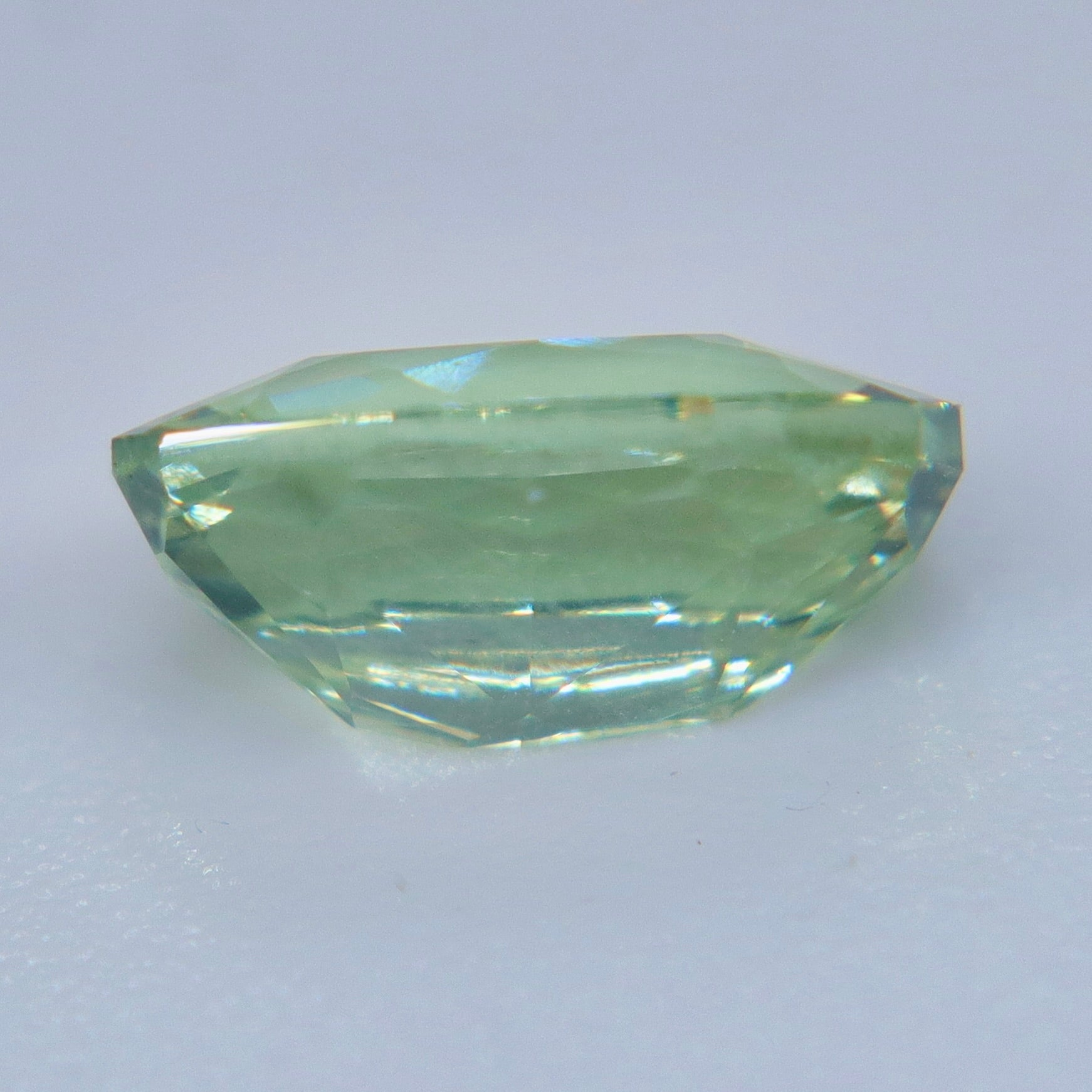 A faceted light green gemstone with a rounded, cushion-like shape, displayed on a plain light gray background.